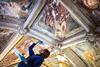 A photo shows a woman wearing a blue lab coat and blue nitrile gloves concentrating on using a brush to touch up a large fading ceiling fresco