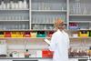 A photo showing a white blonde woman in a white lab coat. She's standing with her back to the camera in front of a lab bench, holding a clipboard on her left hand, inspecting at a wall full of shelves of glassware and other lab equipment