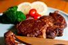 A photograph of a piece of steak on a plate, arranged with some tomatoes, broccoli and slices of lemon. A small piece of the steak has been cut off with a wooden knife that is lying over the side of the plate.