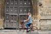 A photo of an large wooden gate featuring several coats of arms. It's the entrance door to one of the colleges at the University of Oxford. A person on a bike is cycling past, the motion blurring the details