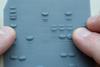 A close-up photo of someone's fingers using a lithophane of a protein electrophoresis gel. The lithophane is a grey-blue flat plastic piece with a number of raised elongated spots.