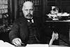 A black and white photo of well-dressed bald white man with a mustache and goatee in a messy office