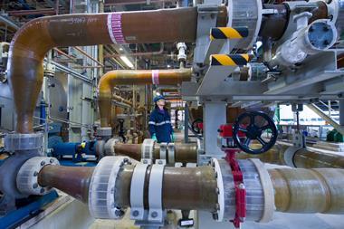 A man wearing safety gear in an industrial chemical plant with large pipes and tanks