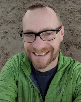 Portrait of Lee Hibbett standing on a beach, looking into the camera and smiling